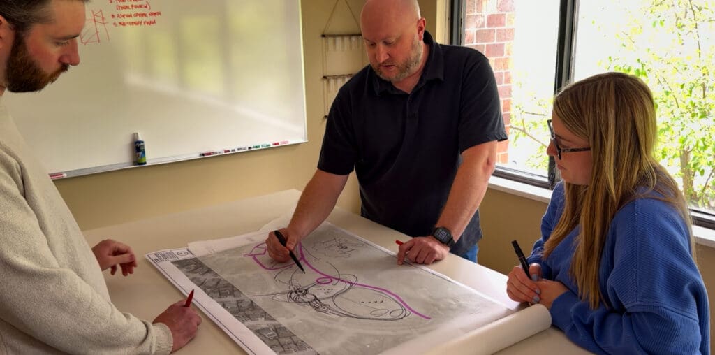Three people lean over a large architectural drawing on a table, each holding a marker as they discuss the design in a bright office. In the background a whiteboard and a window with greenery are visible.