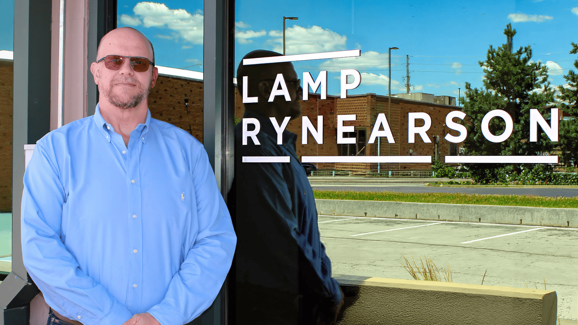 Man in a light blue button-down shirt stands outside a storefront, with a large white LAMP RYNEARSON sign reflected in the glass behind him.